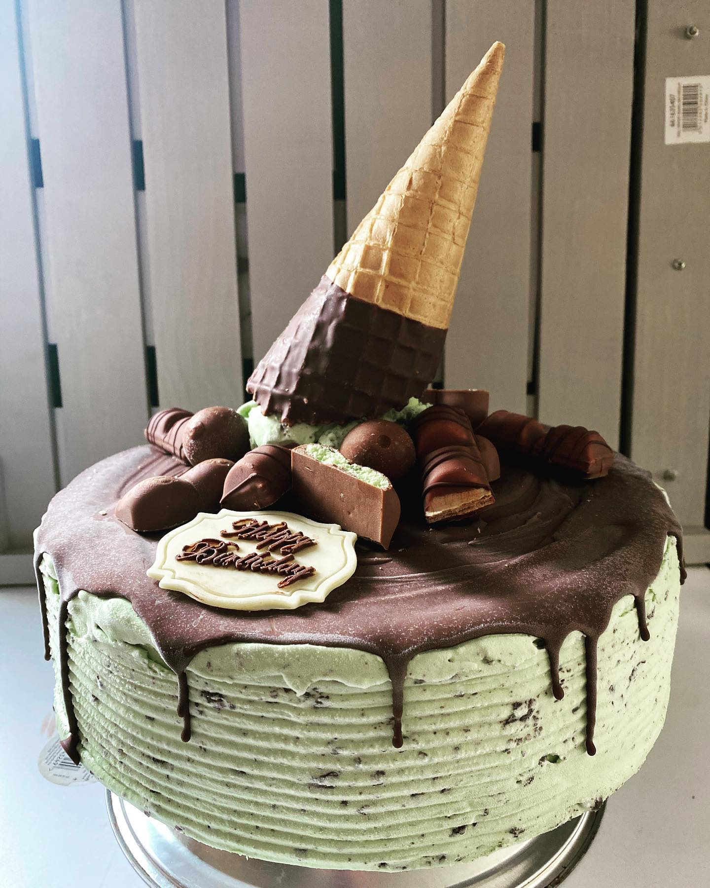 Mint choc ice cream cake topped with bits of mint aero, kinder bueno, a decorated chocolate cone and white chocolate shape with happy birthday written on it 